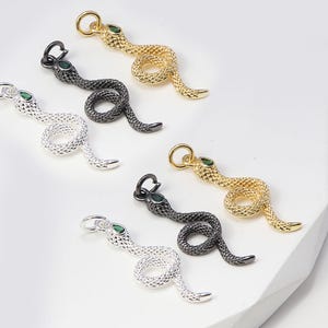 May include: Snake-shaped pendants in silver, gold, and black. Each pendant has a coiled body, a small green gem, and a loop for attachment. The pendants are displayed on a white surface. Ideal for jewellery making.