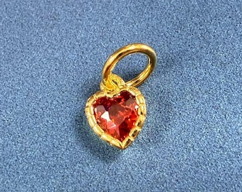 May include: A gold heart charm with a red gemstone in the center. The charm has a loop at the top for attaching to a necklace or bracelet.