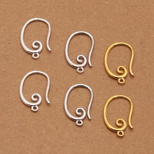 Könnte beinhalten: Sechs Ohrhaken in Silber- und Goldtönen. Die Haken haben ein schlichtes, elegantes Design mit einer Schlaufe oben und einer kleinen Schlaufe unten zum Anbringen von Anhängern oder Perlen. Auf einer hellbraunen Oberfläche präsentiert.