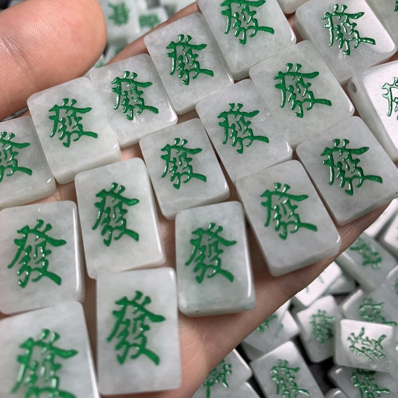 May include: A close-up of a hand holding a pile of white mahjong tiles with green Chinese characters. The tiles are all the same character, which is "發".