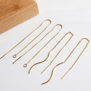 May include: Gold-colored threader earrings in various styles. The earrings feature delicate chains with straight and wavy designs. The earrings are displayed on a white surface with a wooden block in the background.