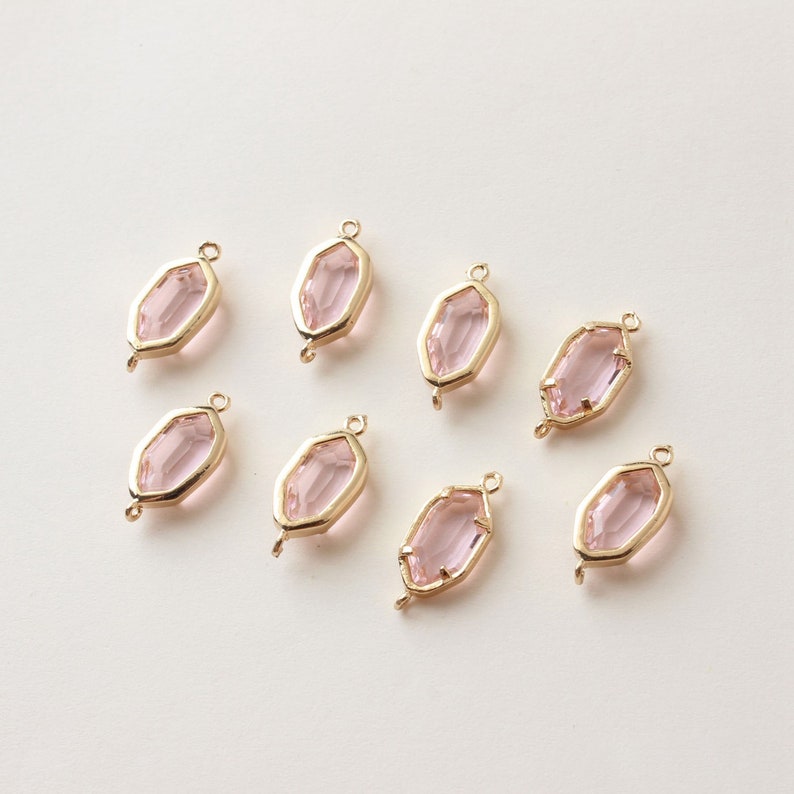 May include: Eight gold-toned, geometric-shaped jewelry charms with a light pink, faceted center. Each charm has a loop at the top and bottom for attaching to other jewelry components. The charms are arranged on a light background.