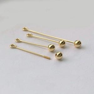 Peut inclure: Une collection de fournitures de bijoux dor&eacute;es, comprenant plusieurs &eacute;pingles droites avec une boule &agrave; une extr&eacute;mit&eacute; et une boucle &agrave; l'autre. Il y a aussi une &eacute;pingle filet&eacute;e et une boule d&eacute;tach&eacute;e. Les fournitures sont dispos&eacute;es sur une surface claire.