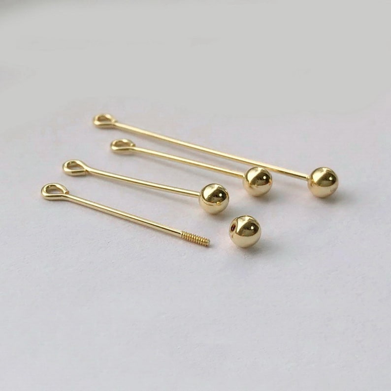 Peut inclure: Une collection de fournitures de bijoux dor&eacute;es, comprenant plusieurs &eacute;pingles droites avec une boule &agrave; une extr&eacute;mit&eacute; et une boucle &agrave; l'autre. Il y a aussi une &eacute;pingle filet&eacute;e et une boule d&eacute;tach&eacute;e. Les fournitures sont dispos&eacute;es sur une surface claire.