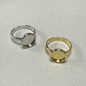 Op de afbeelding: Twee verstelbare ringen met ronde, bezel-gezet, lege, metalen cabochon-instellingen. Een ring is zilver en de andere is goud.