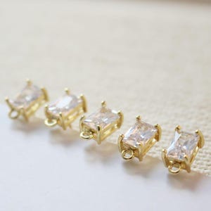 May include: Five gold-coloured rectangular jewellery components with clear, rectangular stones. Each component has a small loop at one end. The components are arranged in a row on a light-coloured surface.