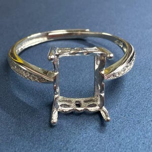 Könnte beinhalten: Ein silberner Ringrohling mit einem rechteckigen Mittelteil für einen Edelstein. Das Band ist mit kleinen, klaren Steinen besetzt. Der Ring wird auf einer blauen Oberfläche präsentiert, um sein aufwendiges Design und sein Anpassungspotenzial hervorzuheben.