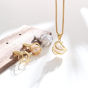 Puede incluir: Un collar dorado con un colgante que presenta una perla blanca encerrada en una jaula de metal en espiral. Dos colgantes adicionales, uno dorado y otro plateado, cada uno con una perla blanca en una jaula similar, descansan cerca.