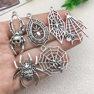 Könnte beinhalten: Eine Sammlung von sechs silberfarbenen Metallanhängern mit Spinnen- und Netzmotiven. Die Anhänger umfassen eine Spinne mit Totenkopf, verschiedene Netzformen mit Spinnen und eine detaillierte Spinne mit strukturiertem Körper.