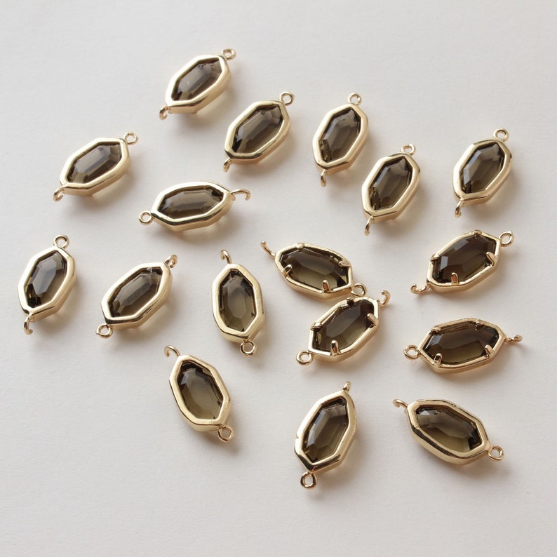 May include: A collection of gold-colored, geometric charms with a smoky quartz-colored center stone. Each charm has a loop on each end for attaching to jewelry. The charms are arranged on a light background.