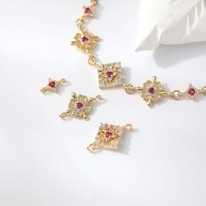 May include: A gold-toned chain necklace featuring repeating square and clover-shaped links, each set with a central red gemstone and surrounded by small clear crystals. Three loose, matching decorative link components are placed nearby.