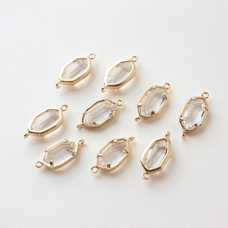 May include: Eight gold-toned, geometric charms with clear, faceted centers. Each charm has a loop at each end for attaching to jewelry. The charms are arranged on a white surface, showcasing their reflective qualities.