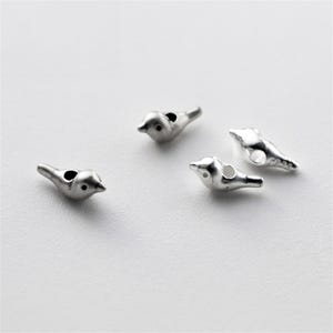 以下が含まれることがあります： 白い表面に散らばる4つの小さな銀色の鳥のチャーム。 各チャームは、小さな穴と詳細なくちばしを備えた鳥のような形をしています。 チャームはおそらく、ジュエリー作りや工芸品用です。