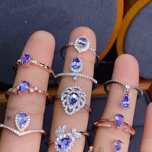 May include: A hand model wearing multiple silver and rose gold rings with teardrop-shaped blue gemstones. The rings are set with smaller clear gemstones.