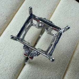 May include: A silver ring setting with a square shaped centre for a gemstone. The setting has a decorative butterfly design with small clear stones on the sides.