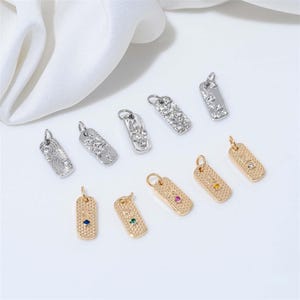 May include: A collection of rectangular pendant charms in gold and silver tones. Some charms feature textured designs, while others have embedded gemstones in various colors. The pendants are designed for jewelry making.