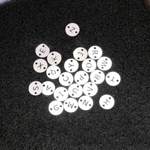 Peut inclure: Un ensemble de petites perles blanches et circulaires avec des lettres de l'alphabet, dispersées sur une surface sombre et texturée. Chaque perle présente une seule lettre dans une police simple. Les lettres visibles incluent S, A, X, Z, C, I, G, W, N, E, J, R, O, B, T et H.