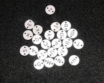 May include: A scattering of small, white, circular alphabet beads on a dark, textured surface. Each bead displays a single letter in a simple font. The letters visible include S, A, X, Z, C, I, G, W, N, E, J, R, O, B, T, and H.