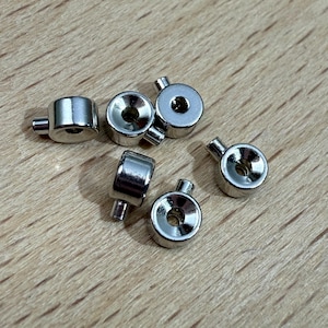 May include: Six small, shiny, silver metal fasteners are scattered on a wooden surface. Each fastener has a cylindrical body, a small post, and a circular recessed area, ideal for various craft projects.
