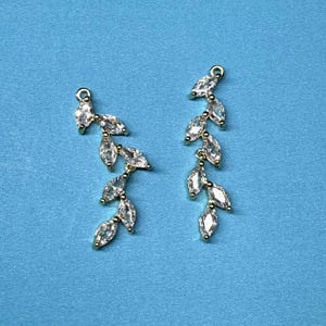 以下が含まれることがあります： 繊細な葉のデザインとクリアなクリスタルアクセントが施された、ゴールドのイヤリングのペアです。
