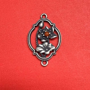 May include: Silver-toned metal pendant with a lotus flower design. The pendant features an oval frame with a detailed lotus flower and bud, and a small orange-brown stone in the center. The pendant has two loops for attaching to a chain or other jewelry.