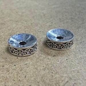 Può includere: Due perline distanziatrici in metallo color argento con un motivo floreale decorativo sui lati. Ogni perlina ha un foro centrale e una parte superiore leggermente concava. Le perline sono su una superficie marrone chiaro.