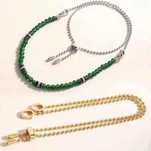 May include: A silver-toned chain bracelet featuring green beads and black and silver spacers, paired with a gold-toned ball chain bracelet. Both bracelets include adjustable sliding mechanisms with metal end caps for a secure fit.