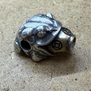 Puede incluir: Un amuleto de rana de metal plateado con un diseño detallado. La rana sostiene un pequeño recipiente redondo de latón con un diseño circular.