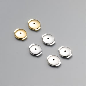 Puede incluir: Un surtido de cierres de pendientes dorados y plateados. Los cierres de los pendientes son redondos con un pequeño agujero en el centro y un cierre de mariposa. Sobre un fondo gris.