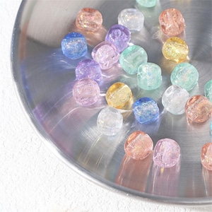 Könnte beinhalten: Eine Sammlung farbenfroher, facettierter Perlen in verschiedenen Pastelltönen, darunter Rosa, Blau, Lila und Gelb. Die Perlen sind auf einer reflektierenden, silberfarbenen Oberfläche verstreut, wodurch ein schimmernder Effekt entsteht. Ideal für Schmuckherstellung und Bastelprojekte.