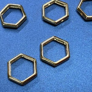 Puede incluir: Primer plano de varios componentes metálicos hexagonales de color dorado. Los componentes están dispuestos sobre una superficie azul. Las formas hexagonales tienen un centro hueco y un acabado brillante.