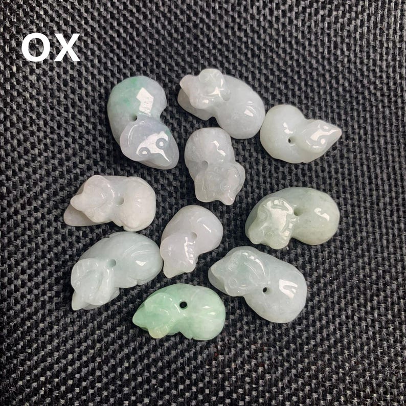 Pode incluir: Uma cole&ccedil;&atilde;o de pingentes de jade esculpidos em forma de boi em v&aacute;rios tons de verde e branco. Cada pingente &eacute; uma pequena escultura detalhada de um boi, com um furo para enfiar. A palavra "OX" est&aacute; no canto superior esquerdo.