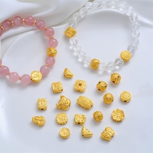 May include: Two beaded bracelets and a collection of loose gold-toned metal beads. One bracelet features smooth pink spherical beads, while the other has faceted clear beads. Both are accented with ornate gold-toned spacers and charms.