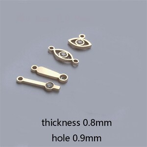 May include: Gold-colored jewelry findings in various shapes, including eye and bar designs, each with a small, clear gemstone. The image includes text indicating a thickness of 0.8mm and a hole size of 0.9mm. These findings are suitable for jewelry making.