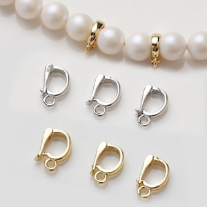 Może przedstawiać: Zbliżenie zapięć do biżuterii, w tym srebrnych i złotych karabińczyków oraz złotych zapięć pierścieniowych. Zapięcia są prezentowane z sznurem białych pereł. Zapięcia są przeznaczone do tworzenia i naprawy biżuterii.