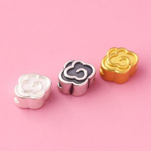 Può includere: Tre ciondoli a forma di fiore in metallo argentato, dorato e nero. I ciondoli hanno un design a spirale.