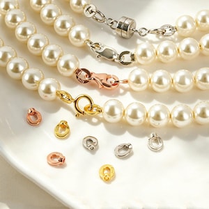 Puede incluir: Tres collares de perlas artificiales de color crema con diferentes cierres metálicos en tonos plata, oro rosa y oro amarillo. Varias anillas metálicas pequeñas y sueltas en acabados a juego están esparcidas cerca de los collares.