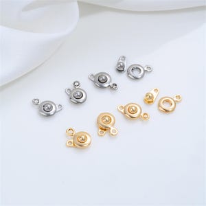 May include: A collection of silver-toned and gold-toned metal snap fasteners arranged in pairs. Each set consists of a circular component with a central stud and a corresponding ring-shaped piece with a small loop for attachment to fabric.