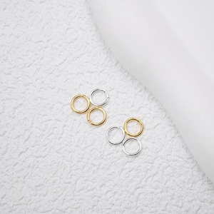 May include: Close-up of six small, circular earrings. Two sets of three earrings are arranged on a textured white surface. One set is gold-colored, and the other is silver-colored. The earrings are simple and minimalist in design.