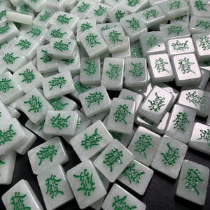 May include: A close-up of a pile of white mahjong tiles with green Chinese characters. The tiles are arranged in a random pattern.