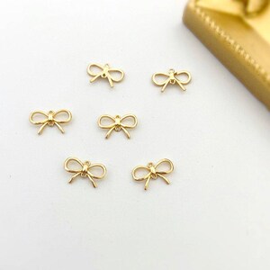May include: Six small gold-toned metal bow charms, each featuring a delicate looped design with a central knot and a small circular jump ring at the top for attachment. These metallic bow-shaped components have a smooth, polished finish.