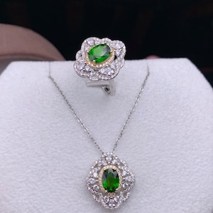 Könnte beinhalten: Ein silberner Ring- und Halskettensatz mit einem grünen ovalen Edelstein, der von kleineren weißen Edelsteinen umgeben ist. Der Ring und der Anhänger haben ein zartes Blumenmuster.