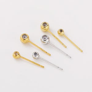 May include: An assortment of gold and silver-coloured jewellery findings. The findings have a ball-shaped top with a small hole and a long, thin post. The jewellery findings are in various sizes and are arranged on a white surface.