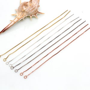 May include: A selection of fine chain necklaces in gold, silver, and copper tones. Each chain has a box-style link design and a small loop at the end. A seashell is visible in the background.