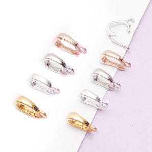 May include: An assortment of jewelry clasps in gold, silver, and rose gold tones. The clasps are in various shapes and sizes, with a small loop at the end for attaching to jewelry. The clasps are displayed on a white and purple background.