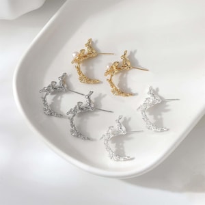 May include: A collection of gold and silver-toned earrings. Each earring features a textured, curved design with a small rabbit and a pearl accent. The earrings are displayed on a white, curved surface.