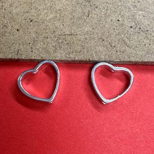 May include: Two silver-colored heart-shaped charms. The charms are open in the center and have a smooth, reflective surface. They are set against a red background with a wooden surface in the upper portion of the image.