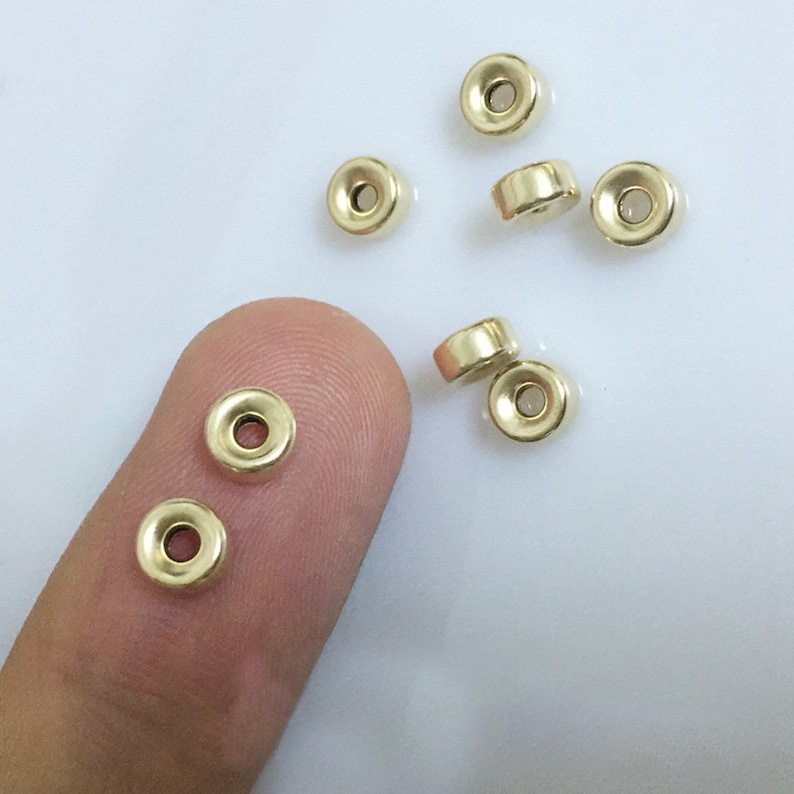 May include: A group of eight gold-colored metal beads with a round shape and a hole in the center. The beads are scattered on a white surface.