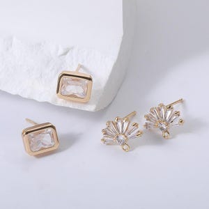May include: Gold-toned stud earrings. Two pairs of earrings are shown. One pair features a rectangular, clear stone set in a gold frame. The other pair has a fan-like design with clear stones and a central, round stone.