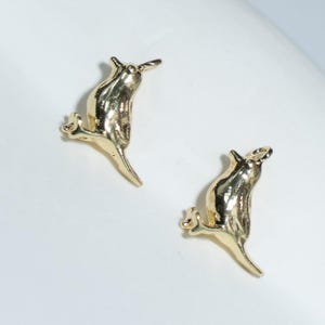 May include: Two gold-colored, stylized rat-shaped stud earrings. The earrings are detailed with small ears, a long tail, and tiny feet. The earrings are set against a white background.